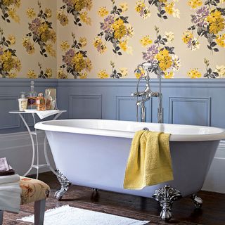 a bathroom with wooden floors, walls where the bottom half is a powder blue and top half a yellow floral wallpaper, and blue and white rolltop bathtub
