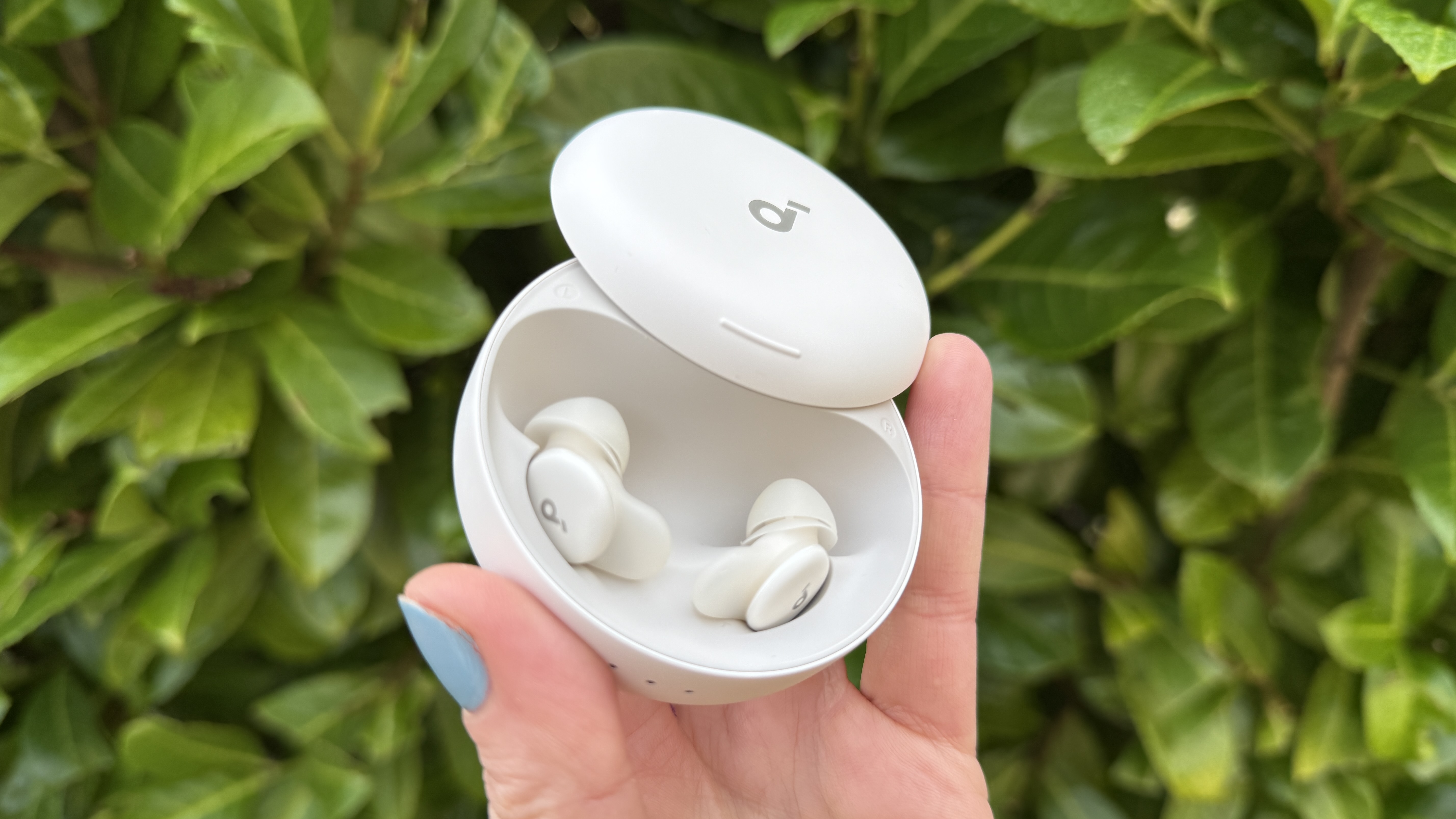 A hand holding the Soundcore Sleep A30 earbuds against a green hedge.