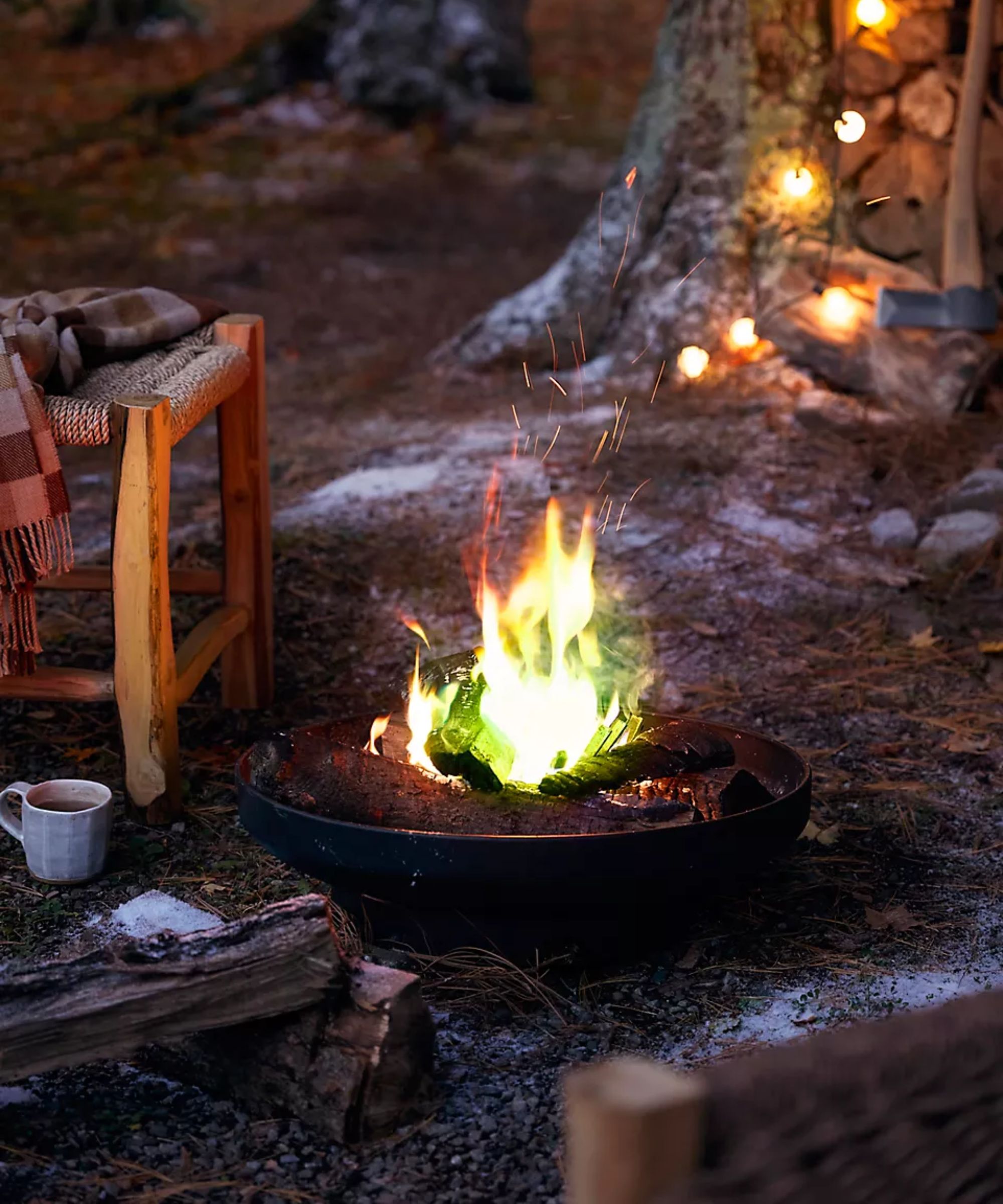 garden area with a large tree strung with fairy lights, with wooden chairs and a burning firepit