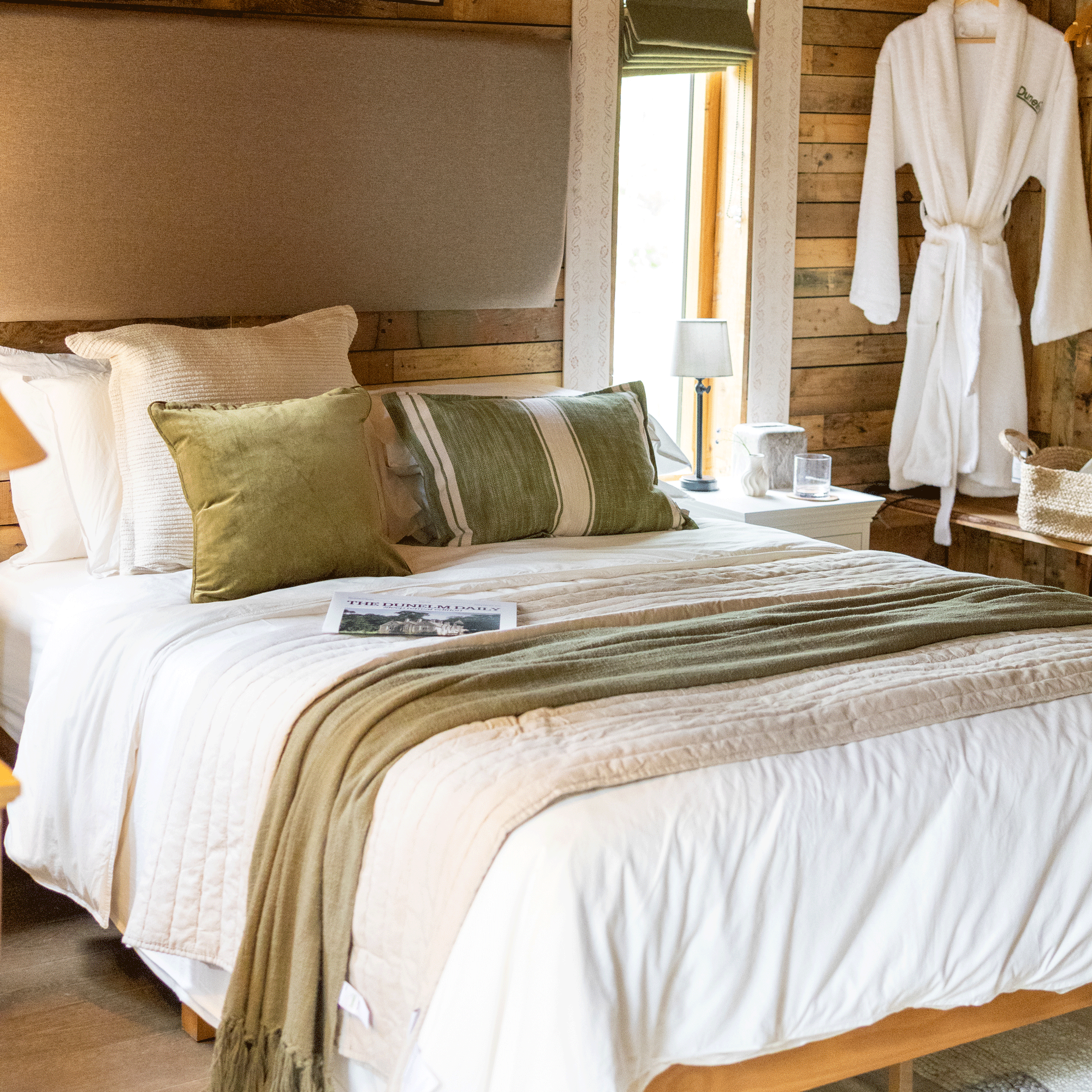a close up of a bed in a wood clad bedroom with layers of white, green and beige bedding