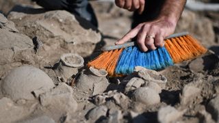 A close-up of a man brushing dust off of ceramics in the ground