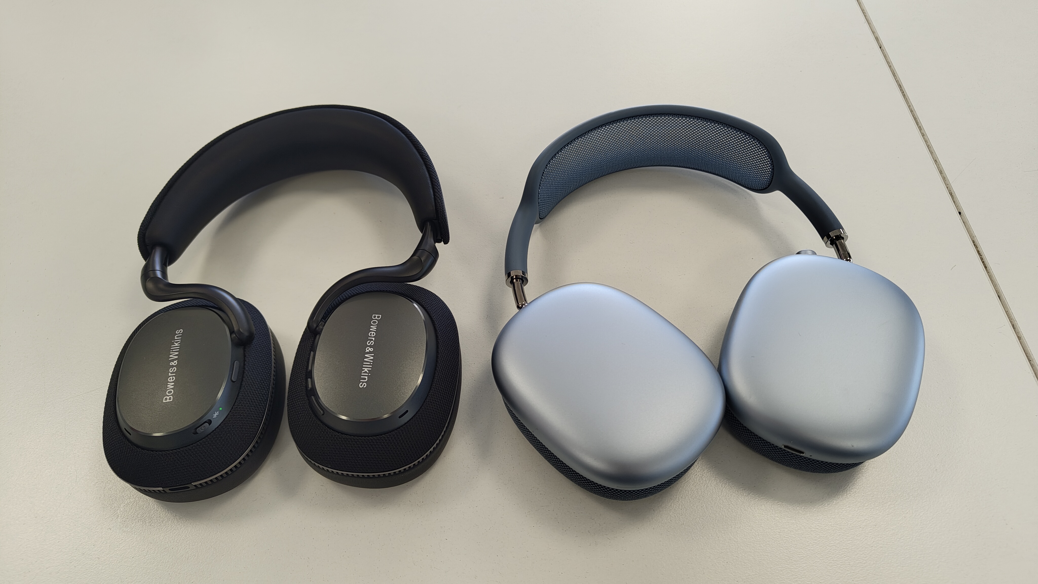 A black pair of B&W Px7 S3 headphones next to a blue pair of Apple AirPods Max headphones. Both are laying flat on a white surface.
