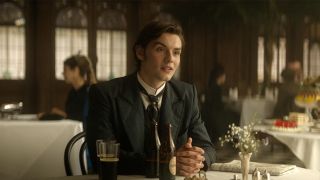 Louis Partridge in a suit, sitting at a table with two bottles of Guinness in front of him in House of Guinness.