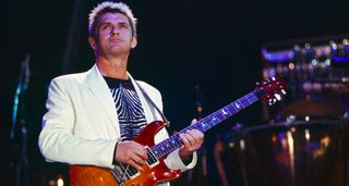 Mike Oldfield wears a white suit jacket and patterned black t-shirt as he plays his PRS Custom 24 onstage.