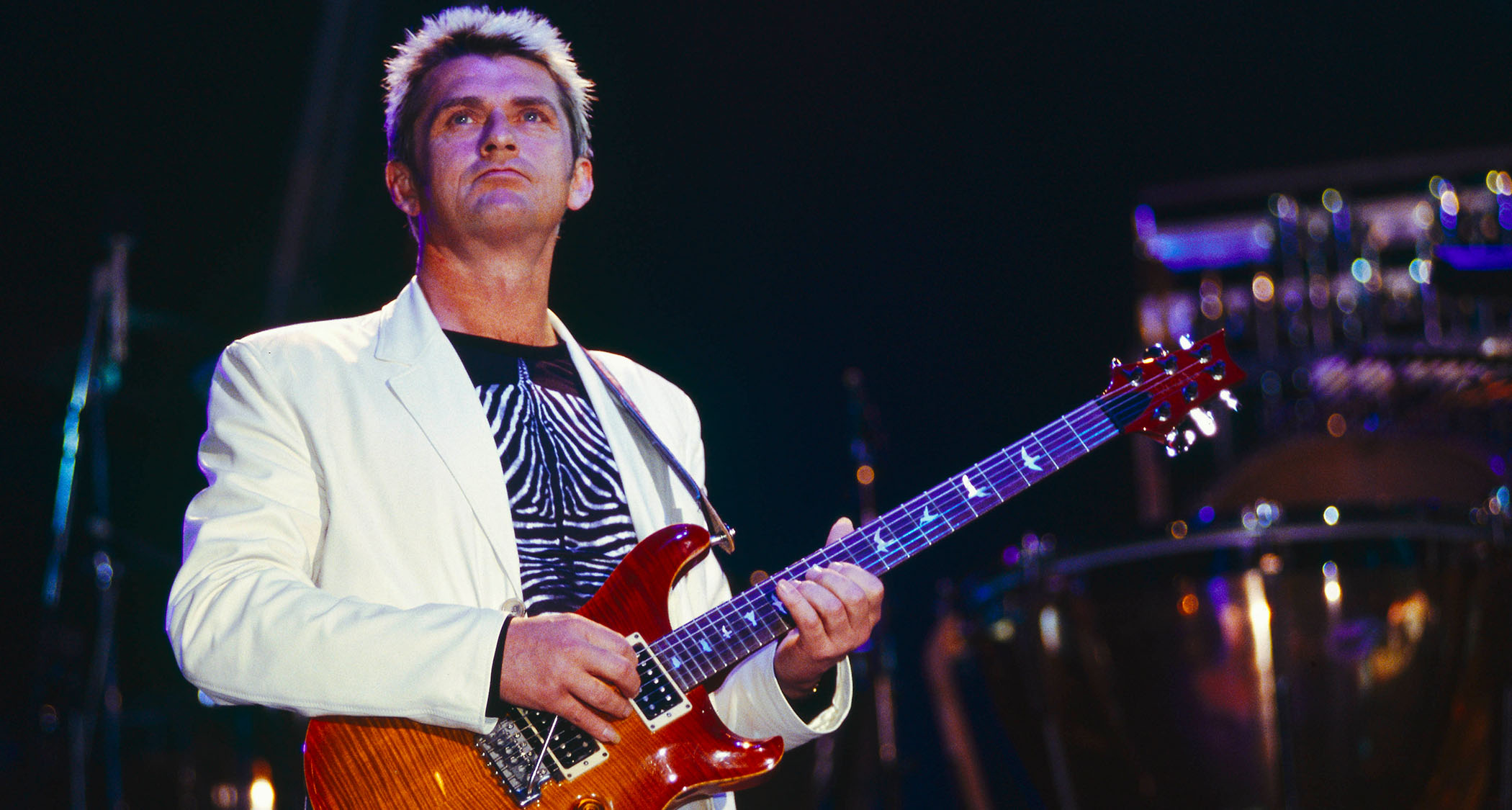 Mike Oldfield wears a white suit jacket and patterned black t-shirt as he plays his PRS Custom 24 onstage.