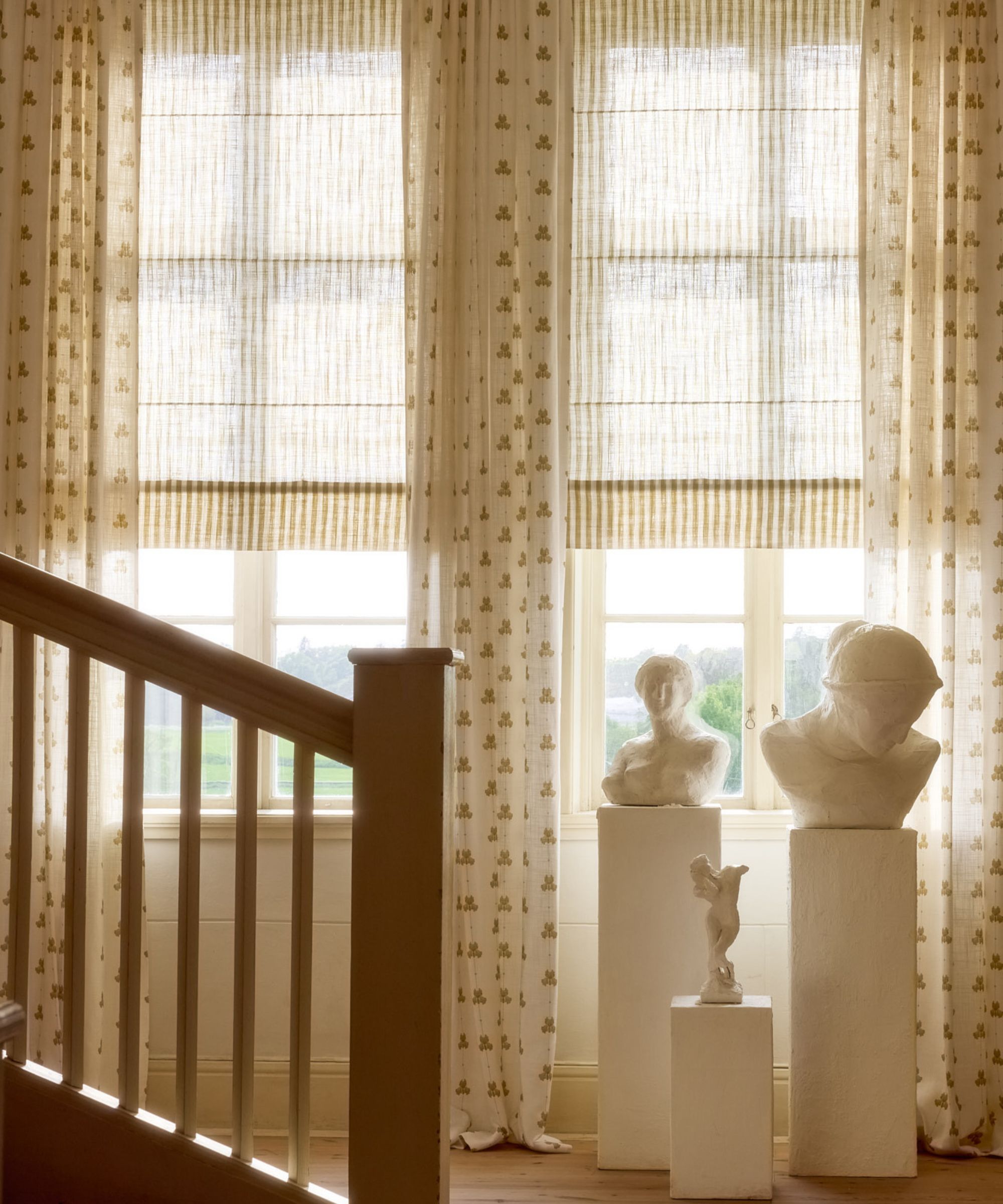 double windows with soft yellow and stripe sheer roman blinds with white sheer drapes with a small yellow floral motif and three plinths with statue sculptures