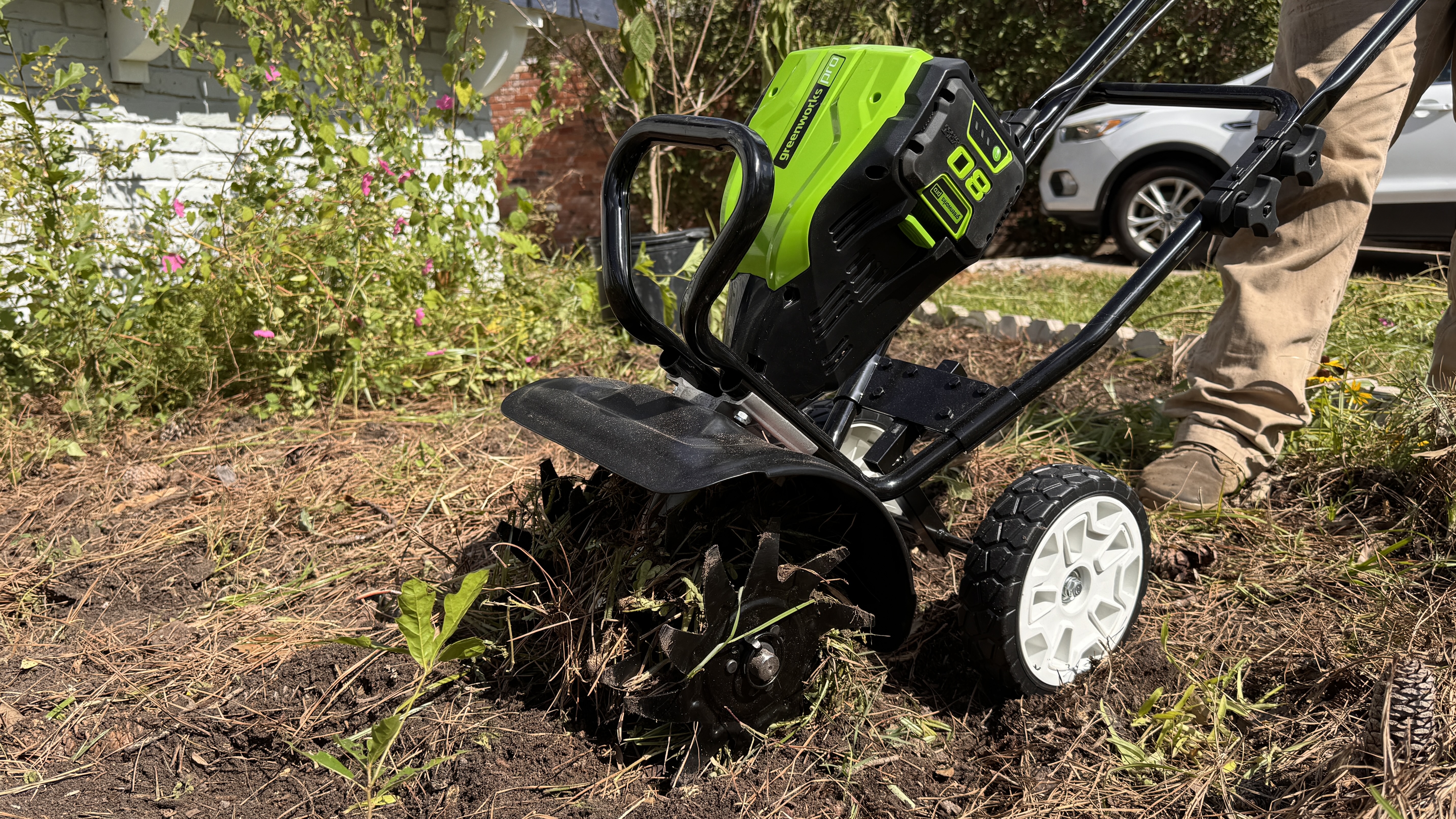 Greenworks tiller used in home garden bed by man in brown pants with white car in the background