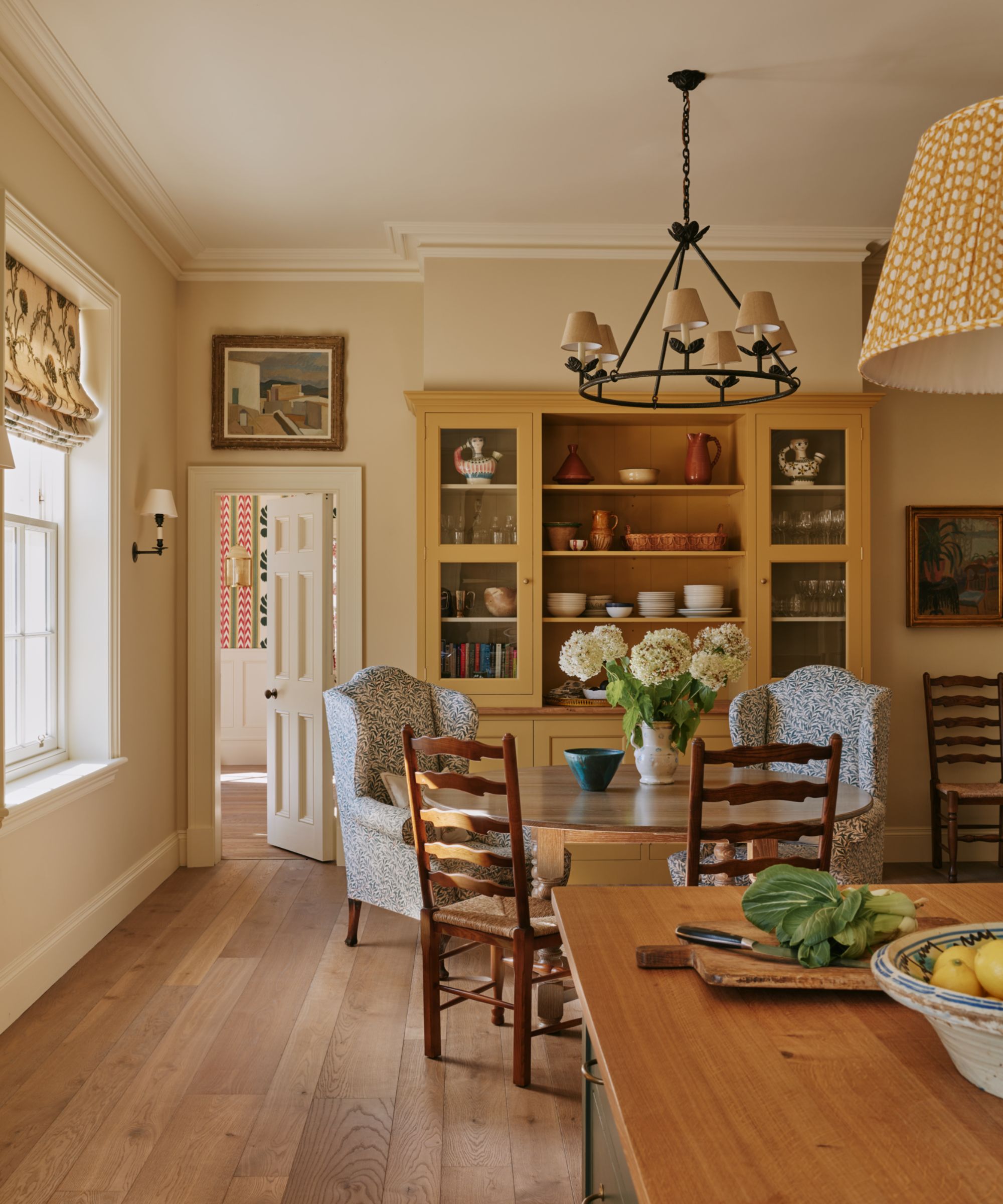 Farmhouse kitchen with wooden floors, a yellow dresser filled with ceramics, a round wooden dining table with two armchairs and dining chairs, and a wooden island with a bowl of fruit on top.