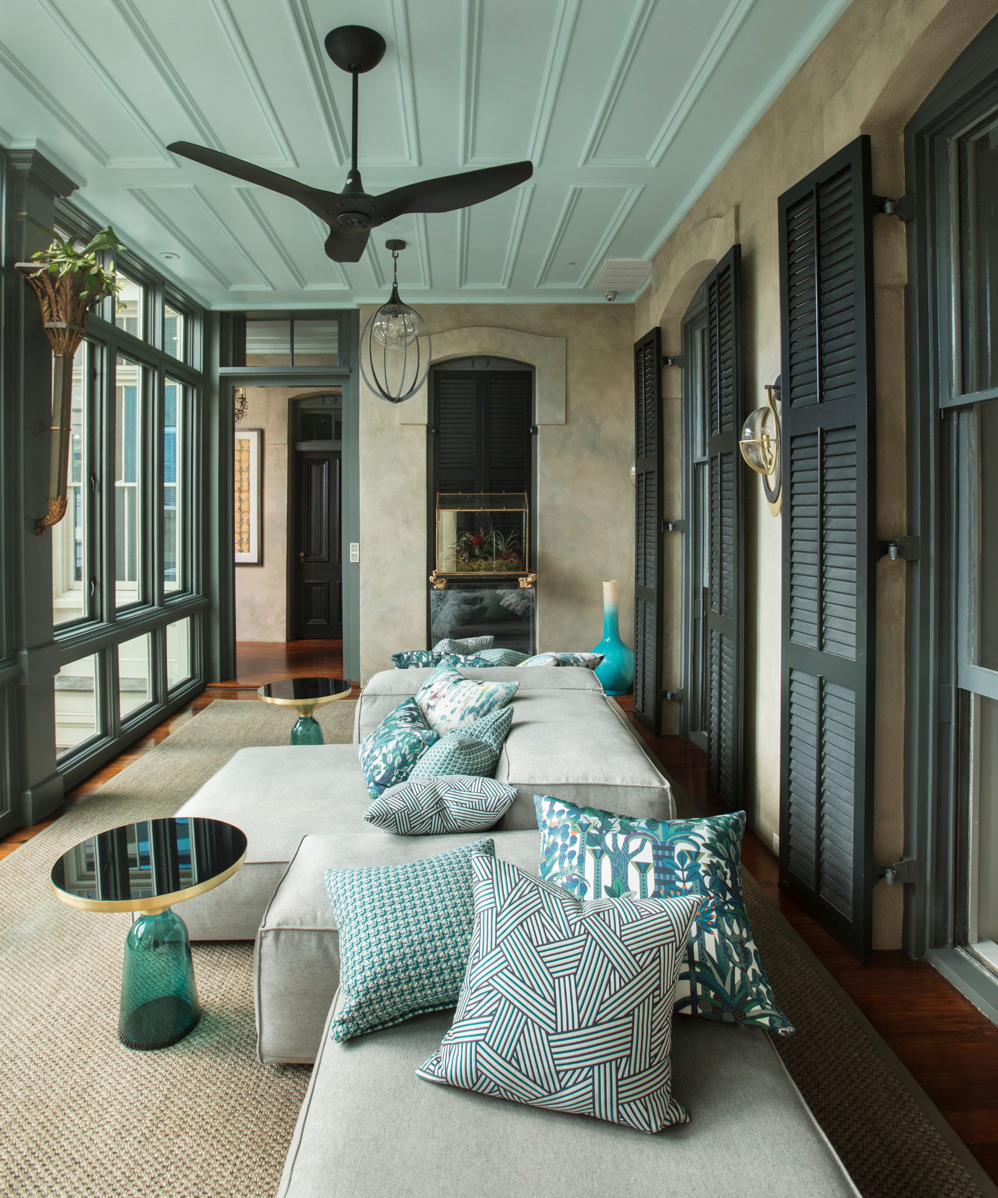 A long sunroom with dark green interior shutters, steel-framed windows, a ceiling fan, neutral modular seating and blue patterned cushions overlooking the outdoors.