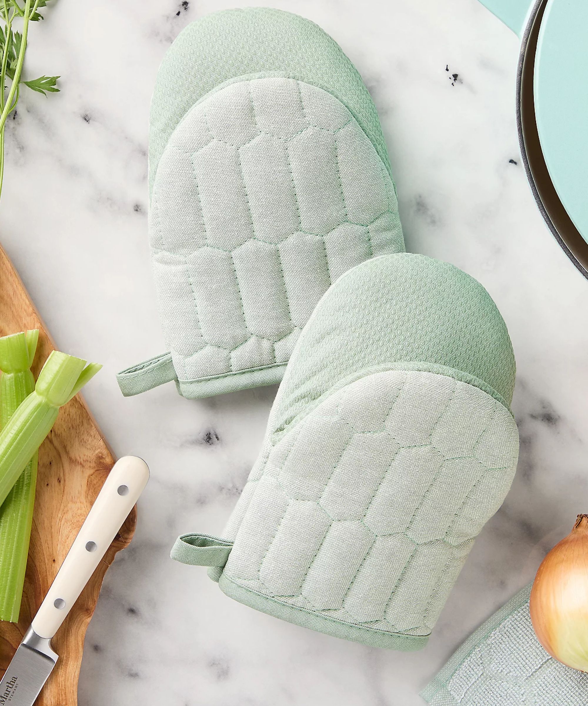 Pale green mini oven mitts on white marble counter next to a wooden chopping board and white knife