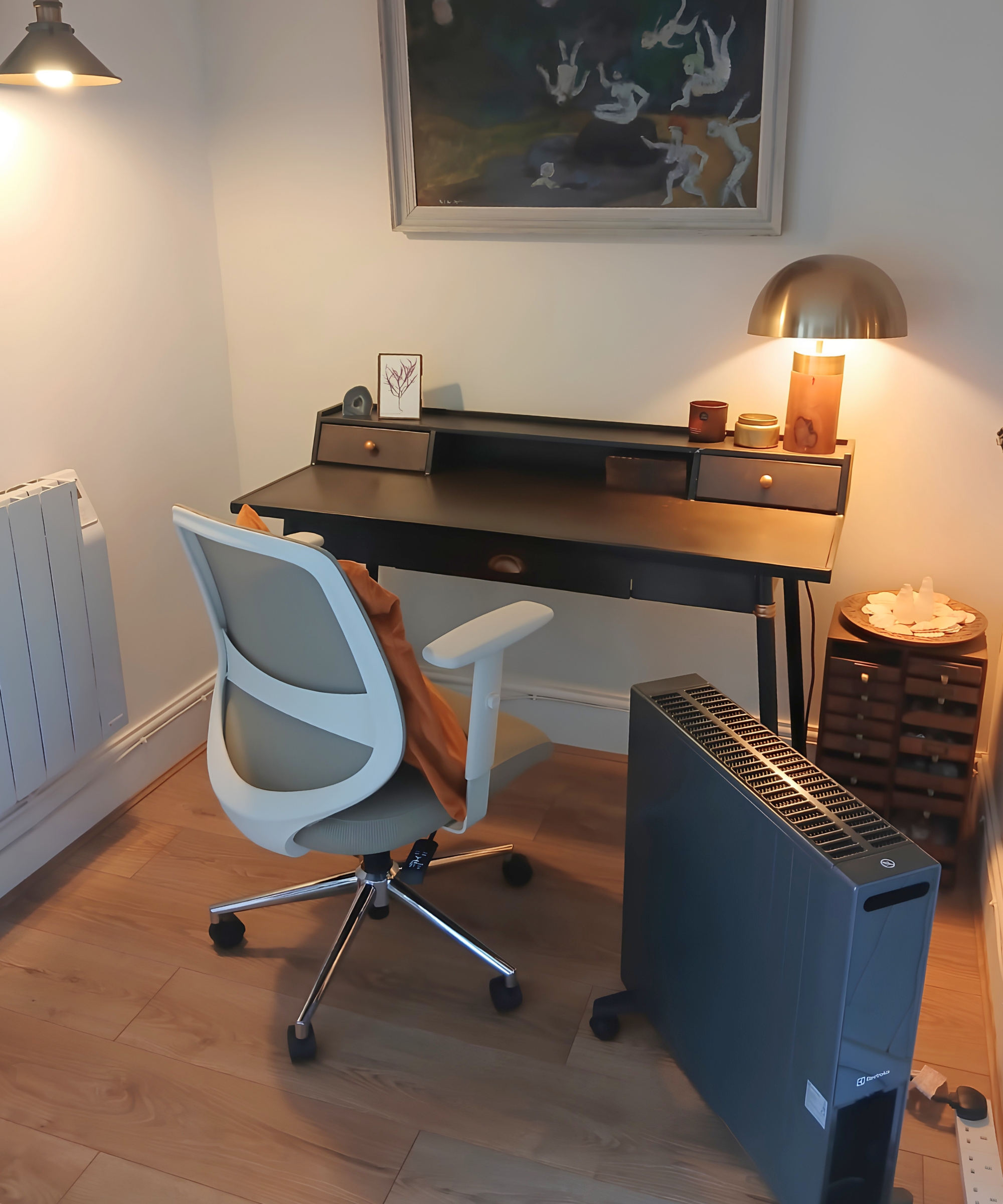 Home office with a mesh desk chair, portable heater, dark metal desk with lamp, and framed artwork above.