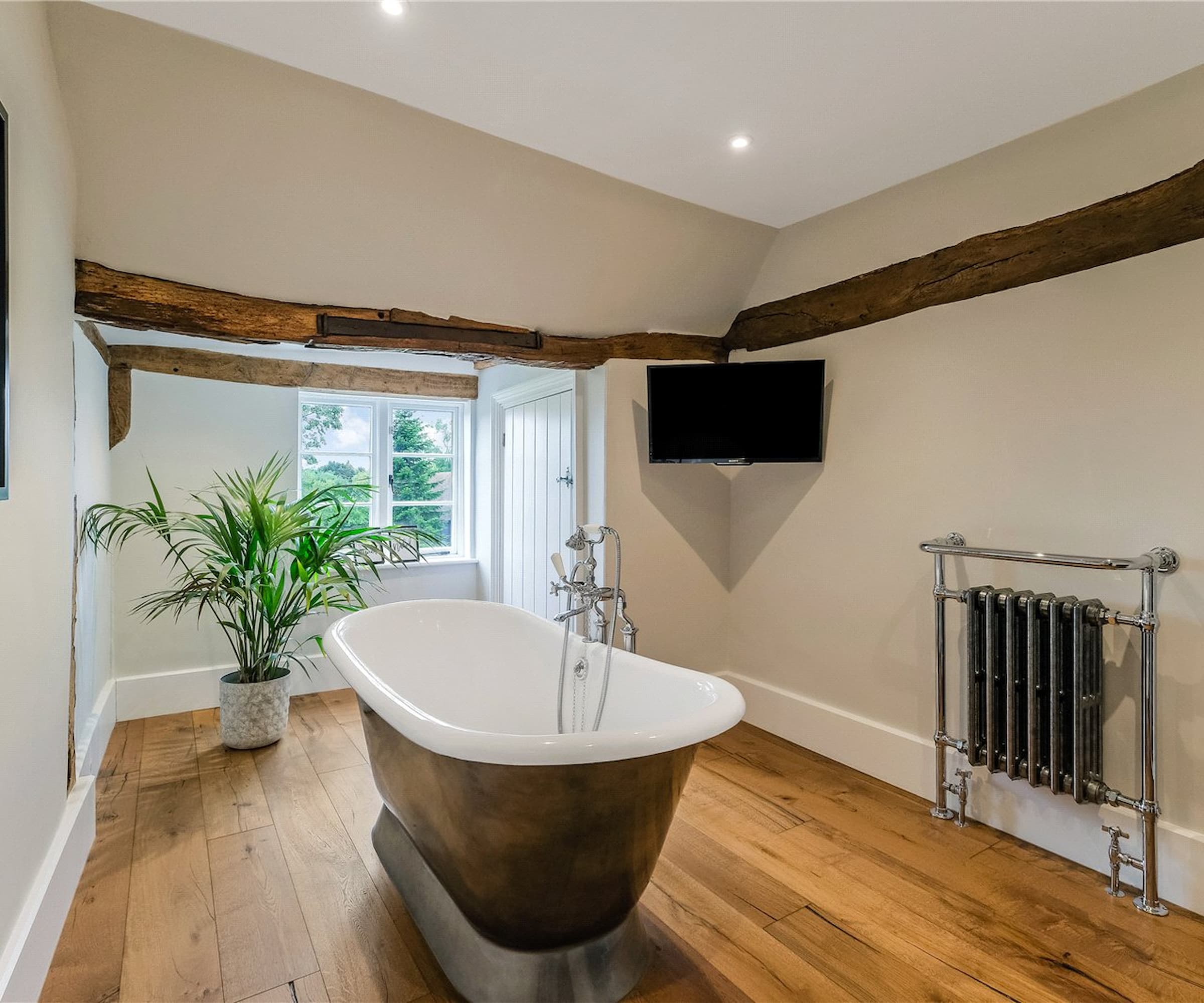 A bathroom with wooden flooring and a metal bath in the middle of the room with oak beams on the roof and a TV in the corner