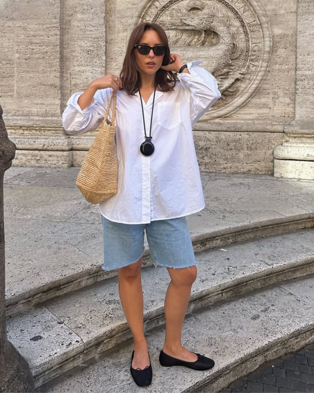 @deborabrosa wears a white linen shirt, denim long shorts, black ballet flats, a raffia bucket bag and cord necklace