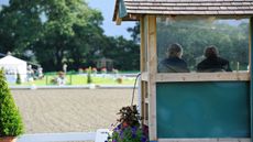 A judges&rsquo; box pictured from behind with a judge and their writer sitting inside facing away from the camera, with an outdoor dressage arena behind.