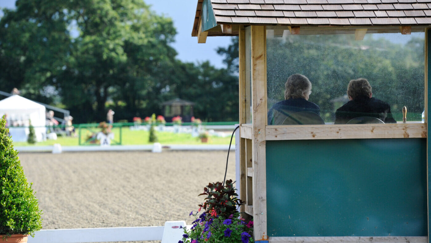A judges&amp;rsquo; box pictured from behind with a judge and their writer sitting inside facing away from the camera, with an outdoor dressage arena behind.