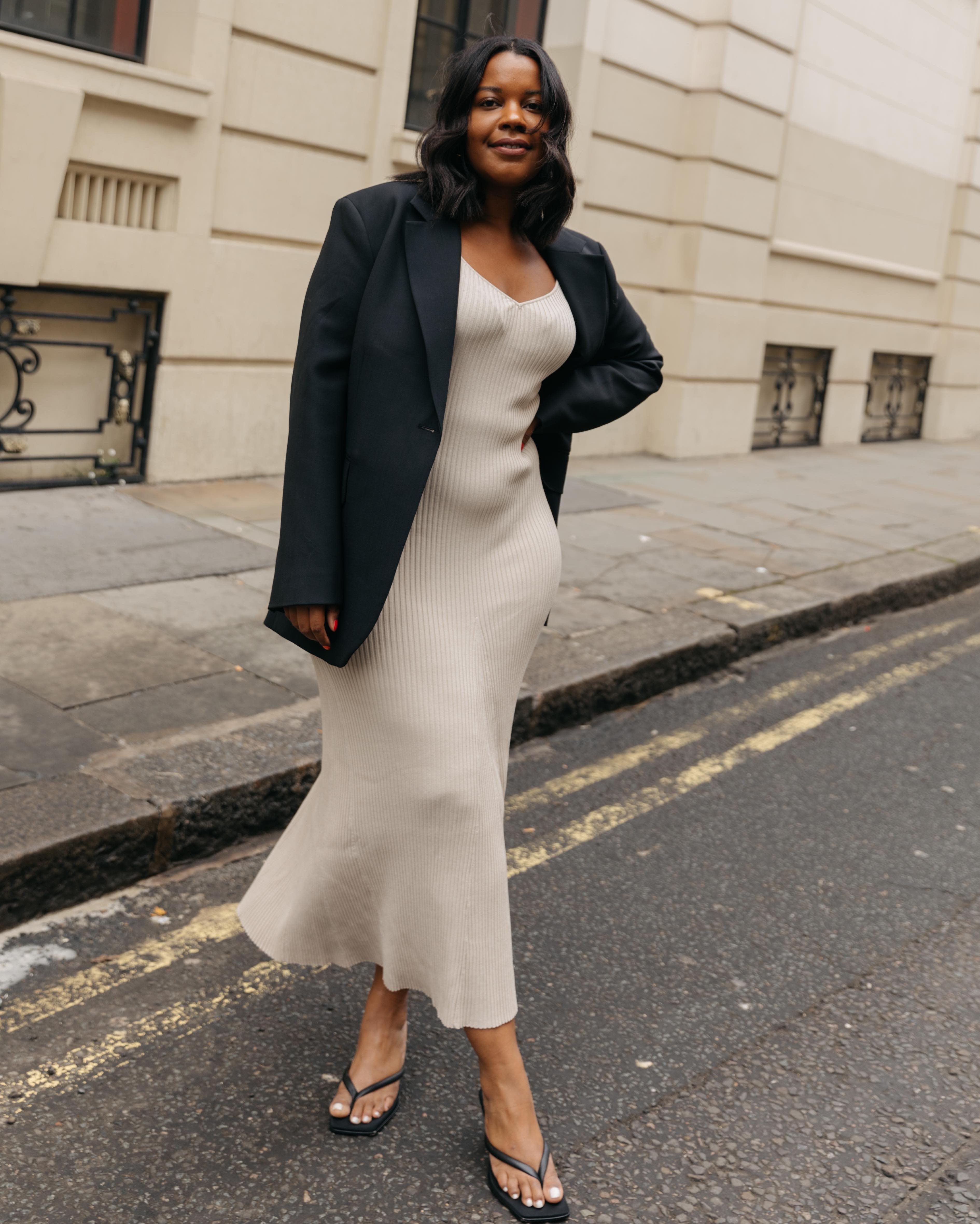 fashion influencer poses on the street in a black blazer, cream ribbed maxi dress, and thong flip flop heel sandals