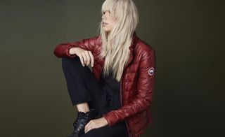 A woman with long white hair wearing a burgundy leather jacket, black pants and black boots with no laces sitting with one knee up.
