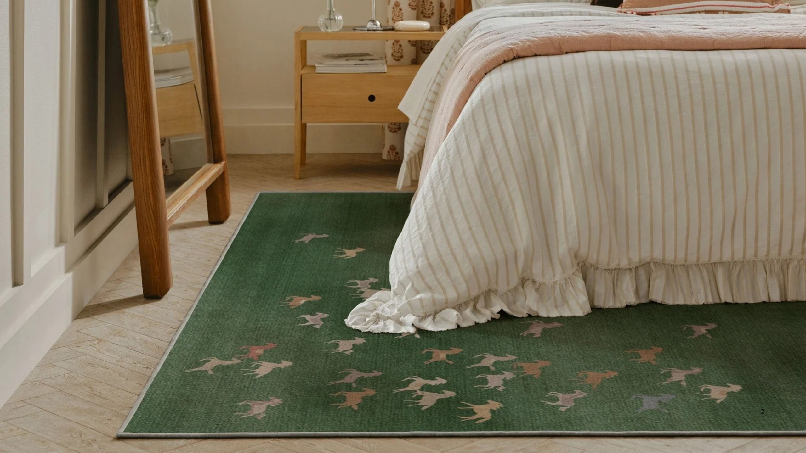 Image of the floor of a bright bedroom that has a dark green rug with small colorful horses on it. There is a bed with white striped bedding, a wooden mirror, and a wooden side table.