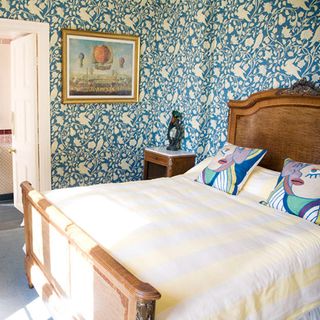 a bedroom with large wooden bed with cushiions on and blue patterned wallpaper and a bedside table