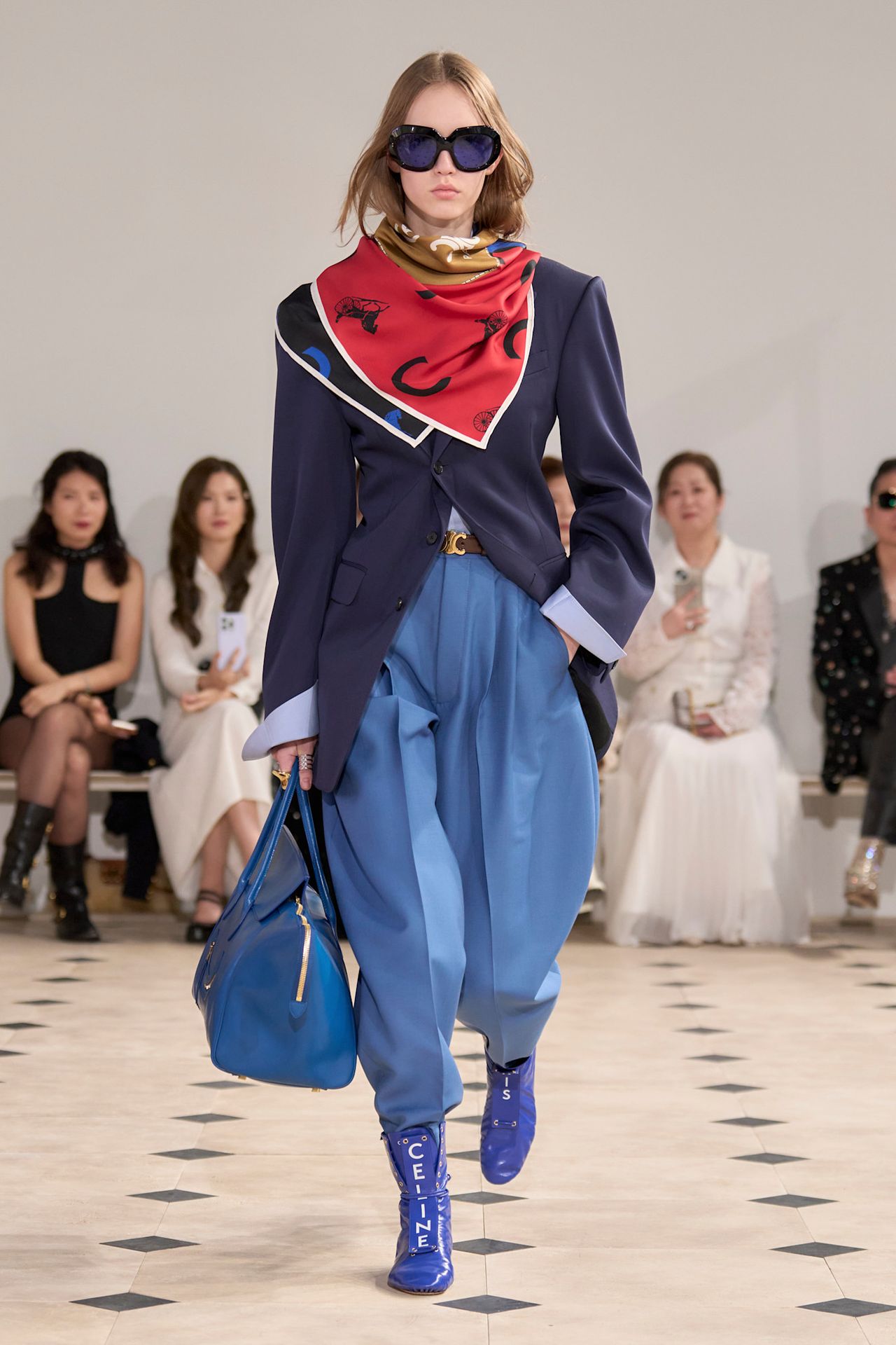 Layering staples for spring 2026 is shown in a photo from Celine's runway show, with a model walking while wearing a printed silk scarf tied around the neck worn with a navy blazer layered on top of a blue button-down shirt styled with blue barrel-leg trousers, blue flat boots, a brown belt, a bright blue bowling bag, and blue tinted lens sunglasses