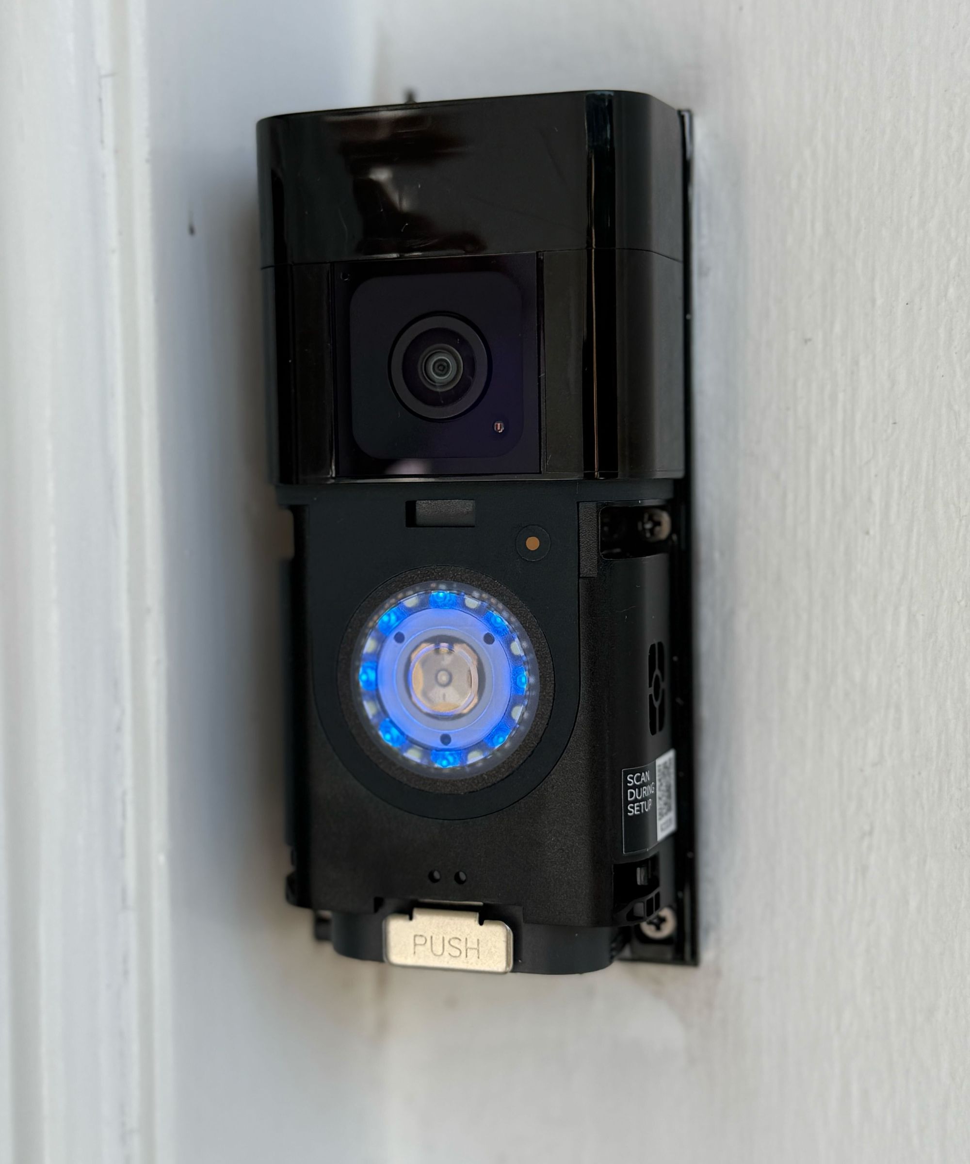 A black Ring Battery Doorbell Plus with the faceplate removed, mounted on a white wall
