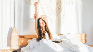 A woman with long dark hair sits up in bed with her arms stretched in the air as sunlight streams in through her open curtains