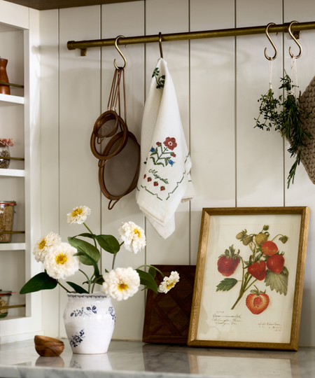 a kitchen countertop with artwork, a vase of flowers, and a pot rack with kitchen necessities hanging