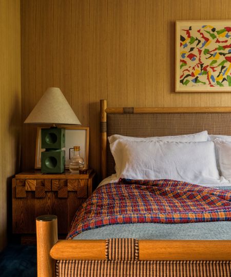 Bedroom with white-oak paneled walls, a striped upholstered bed, white linen, colorful checked quilt, green lamp and abstract art.