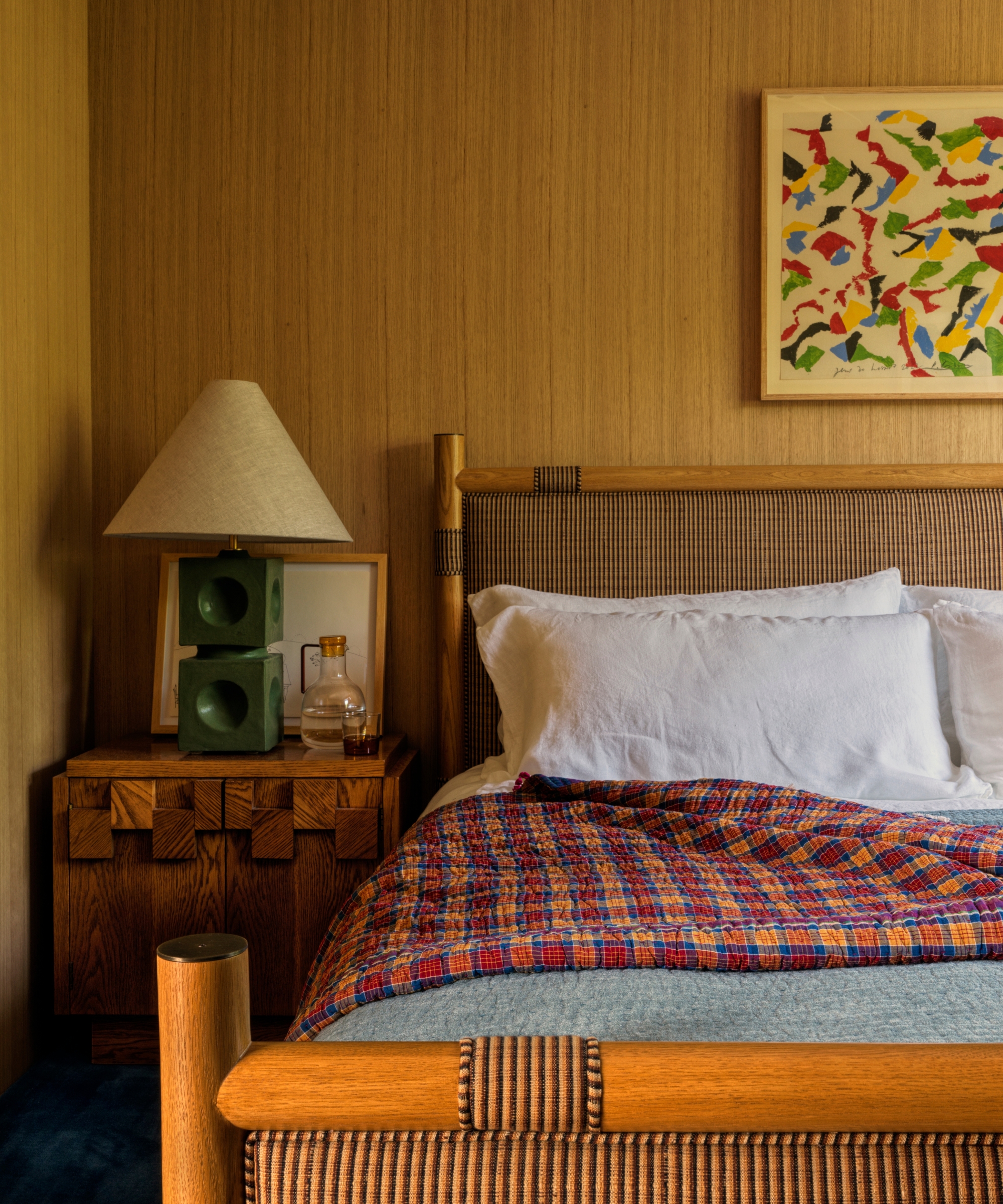 Bedroom with white-oak paneled walls, a striped upholstered bed, white linen, colorful checked quilt, green lamp and abstract art.