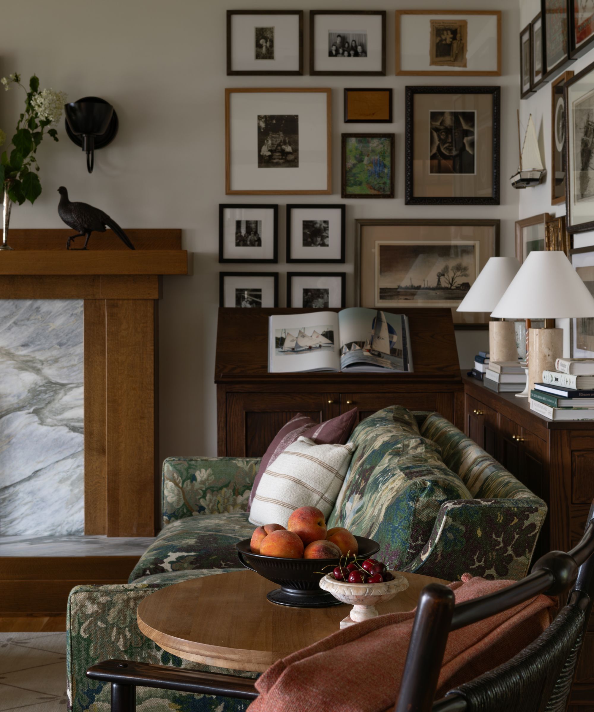 Cosy living room with floral sofa, wooden round side table, mahogany console table with table lamps and stacks of books, marble fireplace, and gallery wall of vintage art