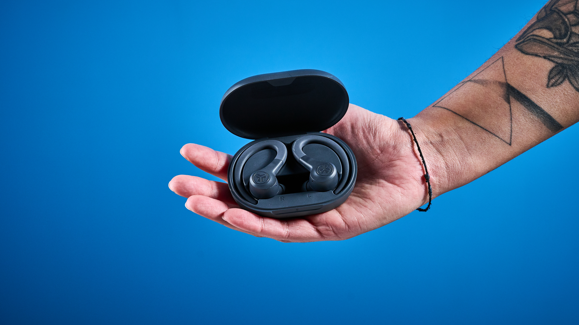 the jlab go sport+ earbuds photographed against the blue tom's guide background, showing charging case and wearing style