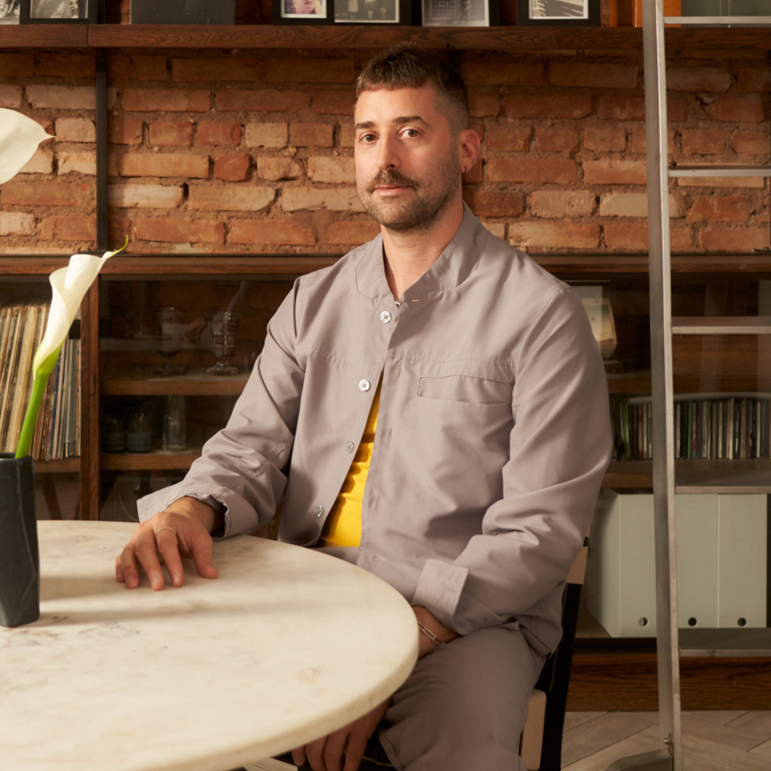 A headshot of a man with short brown hair and a moustache in a gray co-ord with a yellow t-shirt, at a marble dining table with a vase of calla lilies in front of a bookshelf with a steel ladder