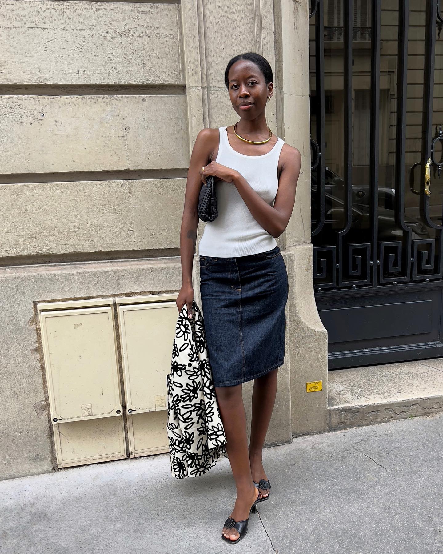 Influencer Sylvie Mus poses on a sidewalk in Paris wearing a collar necklace, white knit tank top, black clutch bag, knee-length denim skirt, and black mule sandals.