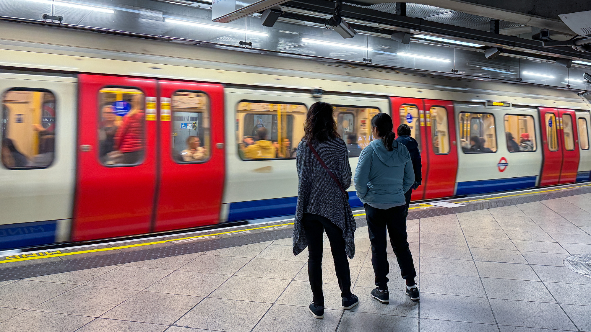 Two women stand on the London underground platform, watching a train pull into the station