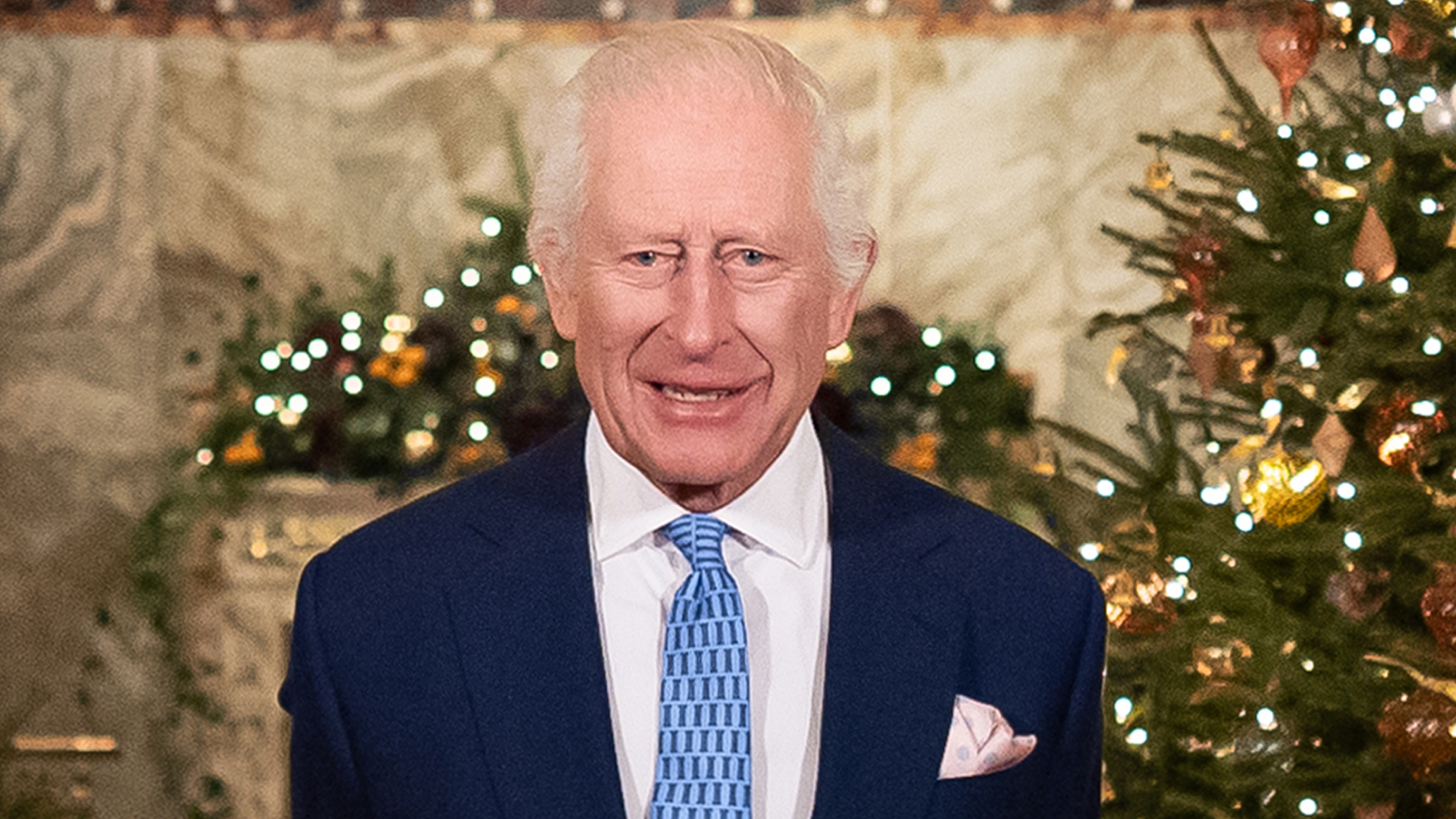 King Charles standing in front of a lit Christmas tree in a blue suit and tie