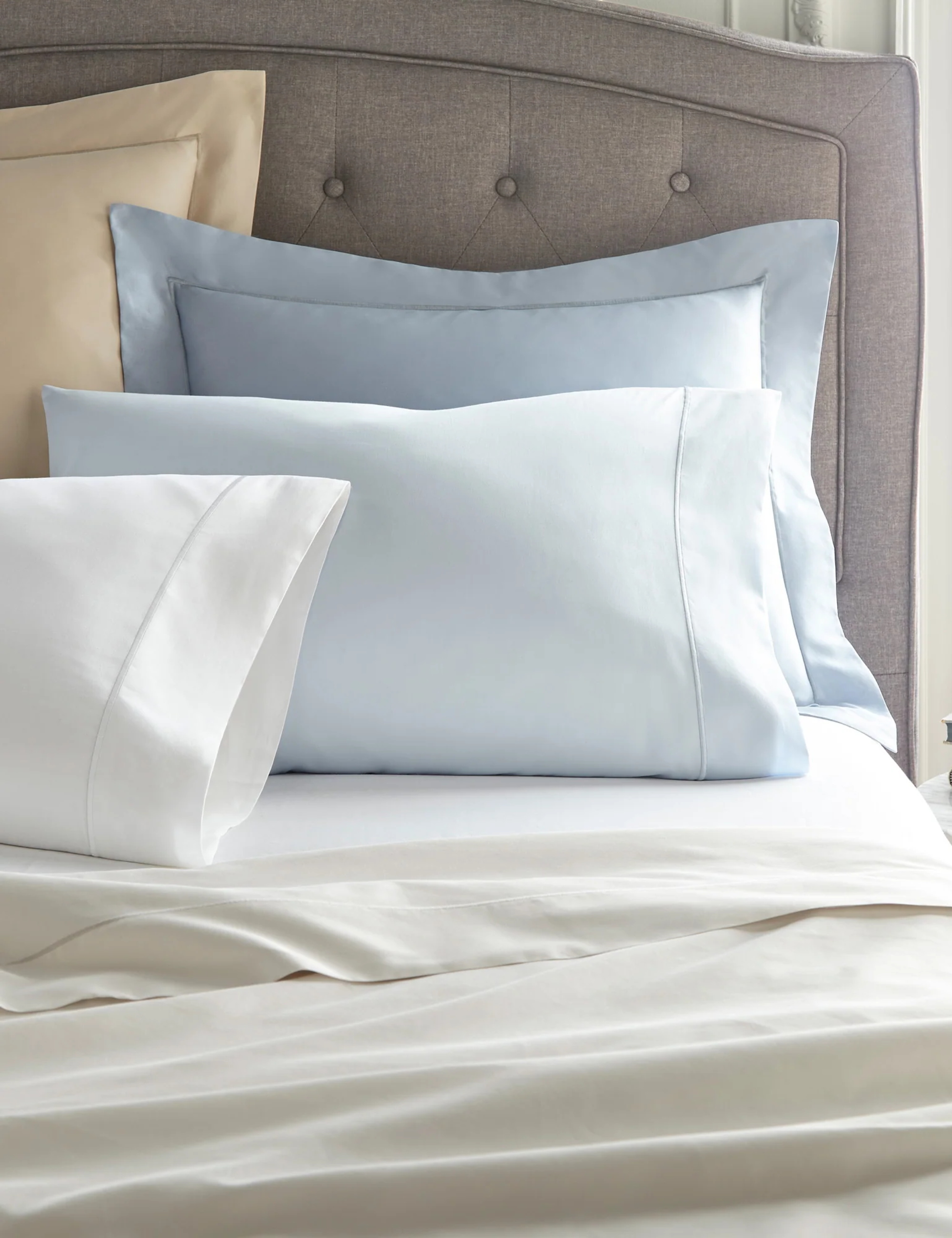 Peacock Alley sateen sheets in various shades of blue and cream styled atop of a bed with a tufted headboard