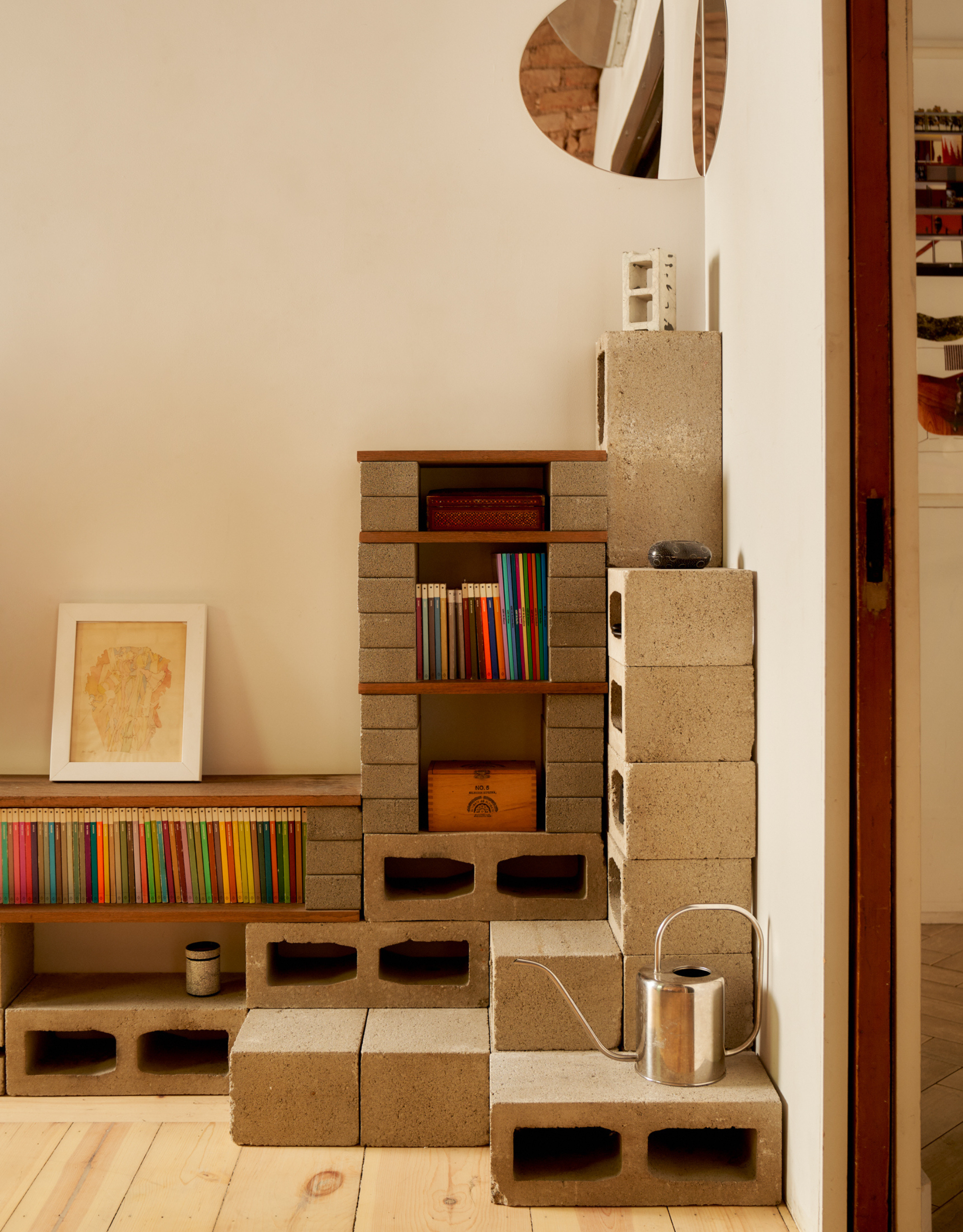 A living room corner with cinder blocks and wooden slabs making up storage nooks filled with books, and a steel watering can propped to the side