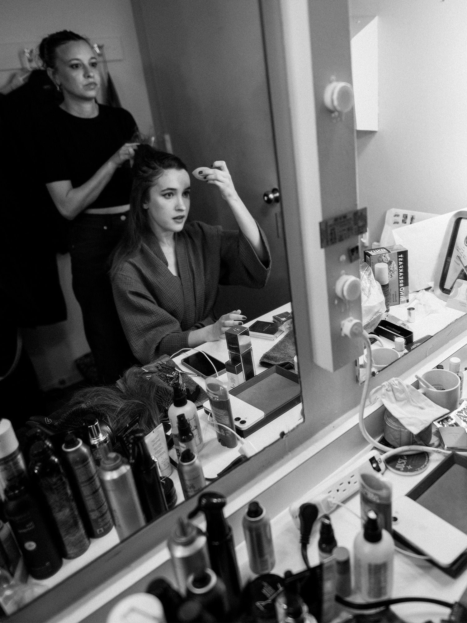 Gracie Lawrence getting ready for the Tony Awards backstage at her musical Just in Time. She is sitting at a glam station looking into a mirror doing her makeup while her hairstylist preps her hair.