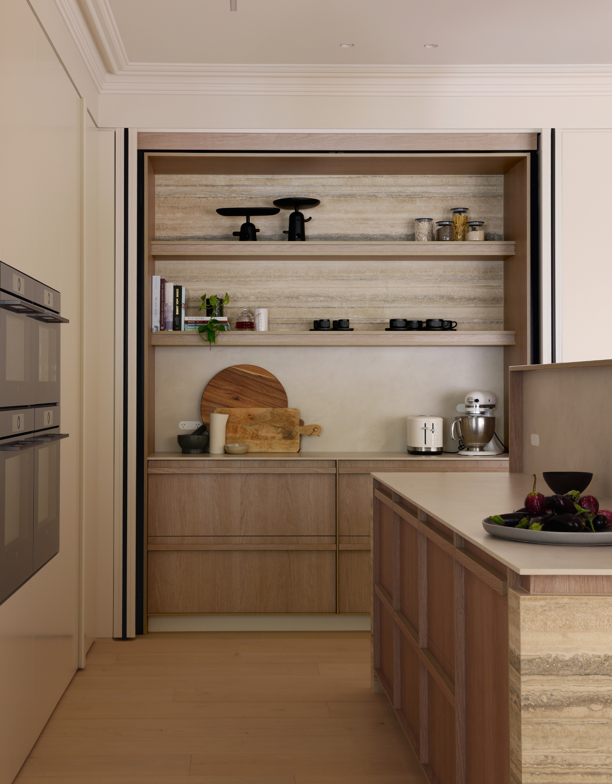 A bright minimalist kitchen with a hidden storage space holding cutting boards, toaster, a blender, a mortar and pestle, and cup seats