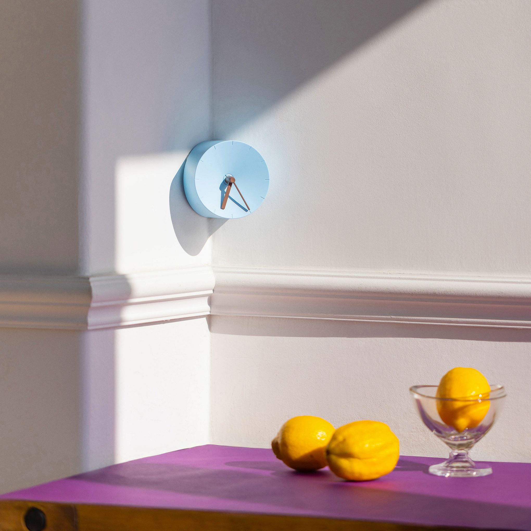 A powder blue circle clock hangs in a corner above a purple table with lemons.