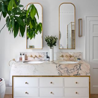 White painted bathroom with marble double sink vanity and storage, gold rimmed mirrors hung on wall, and houseplants displayed