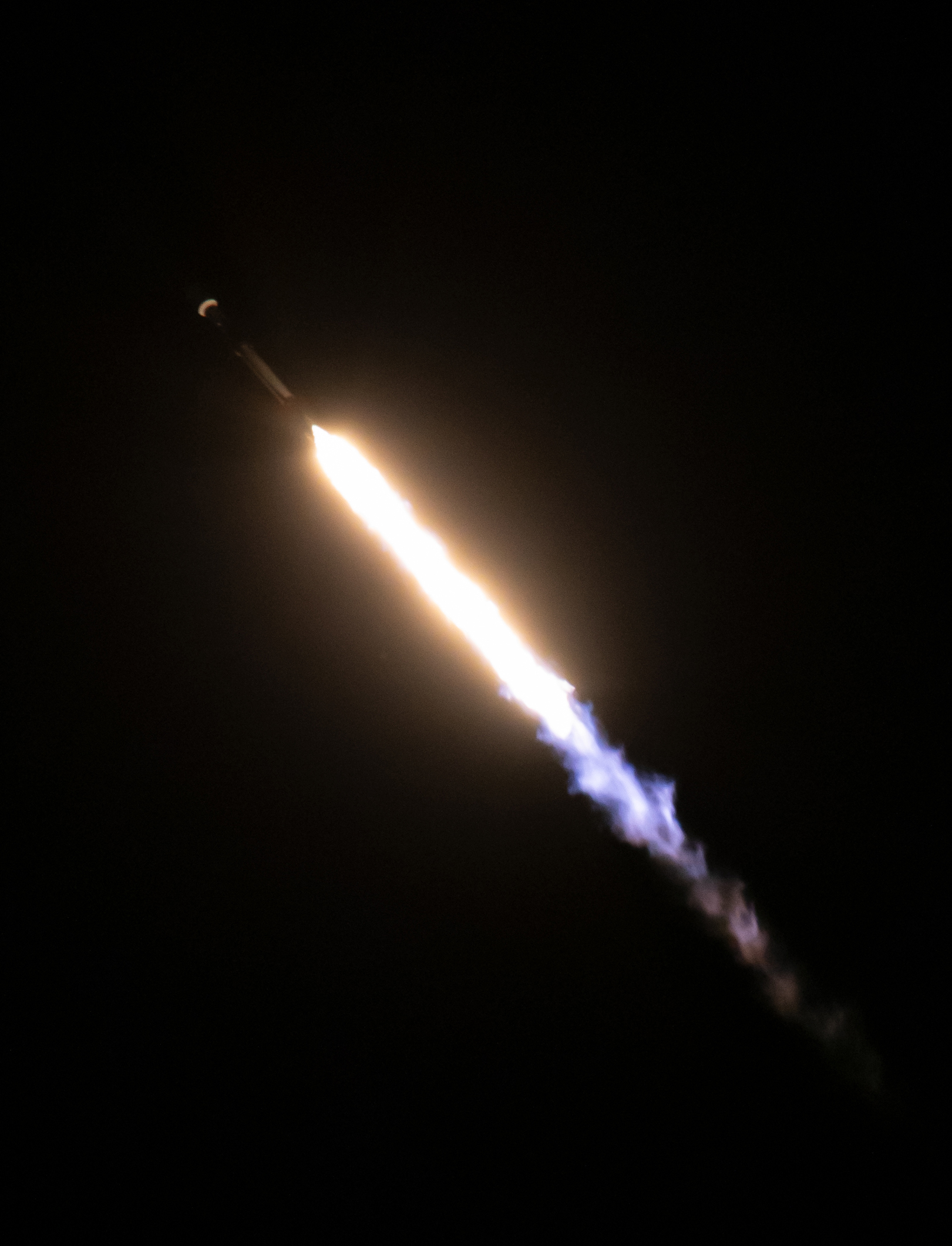 The faint image of a rocket is seen in the shadows of the black night sky as a yellow-white fire bursts from its end, fading to shades of blue and purple as the rocket ascends.