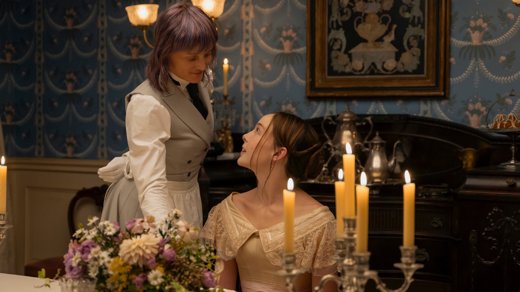 Maggie serves Lizzie a meal set on a candle-lit dining room table. There's a flower arrangement on the table and both women are smiling. 