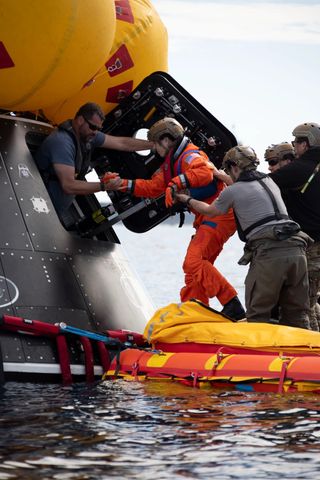 an astronaut stepping into a spacecraft floating on a raft in the ocean. people around her are physically holding her up due to the rough waters