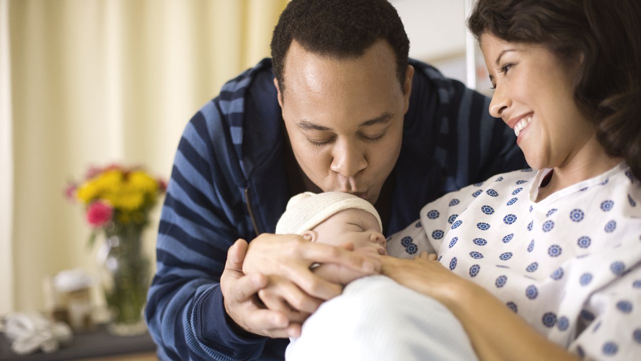 New parents gaze lovingly at their newborn while Dad kisses the baby&#039;s head and Mom looks on in her hospital bed.