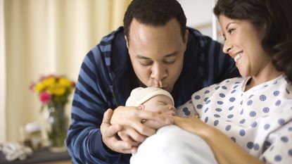 New parents gaze lovingly at their newborn while Dad kisses the baby's head and Mom looks on in her hospital bed.