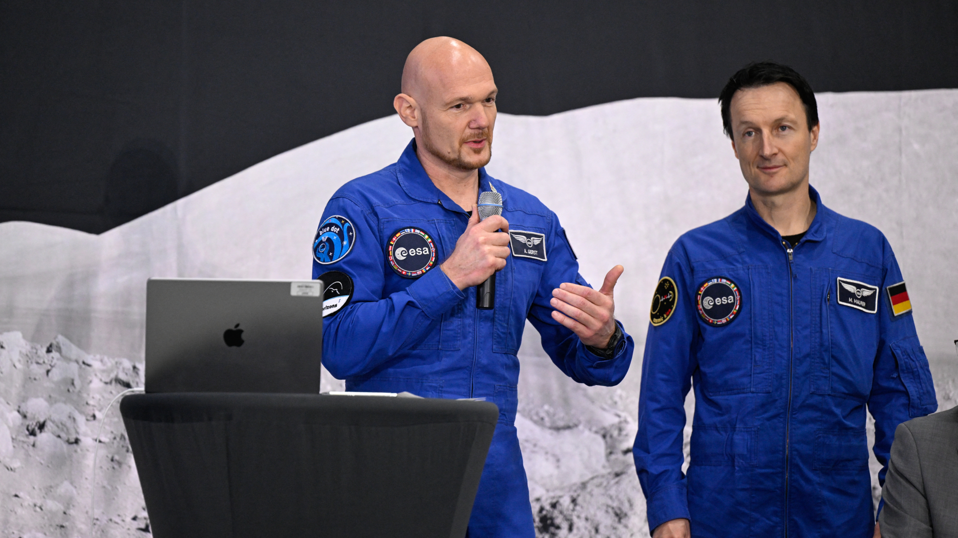 Two men stand next to a podium wearing blue space suits