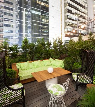 Duddell’s, Hong Kong, China. An outside seating area with a wooden sofa, two black wire chairs, a white wire chair, a rectangular coffee table and potted plants with a view of buildings behind it.