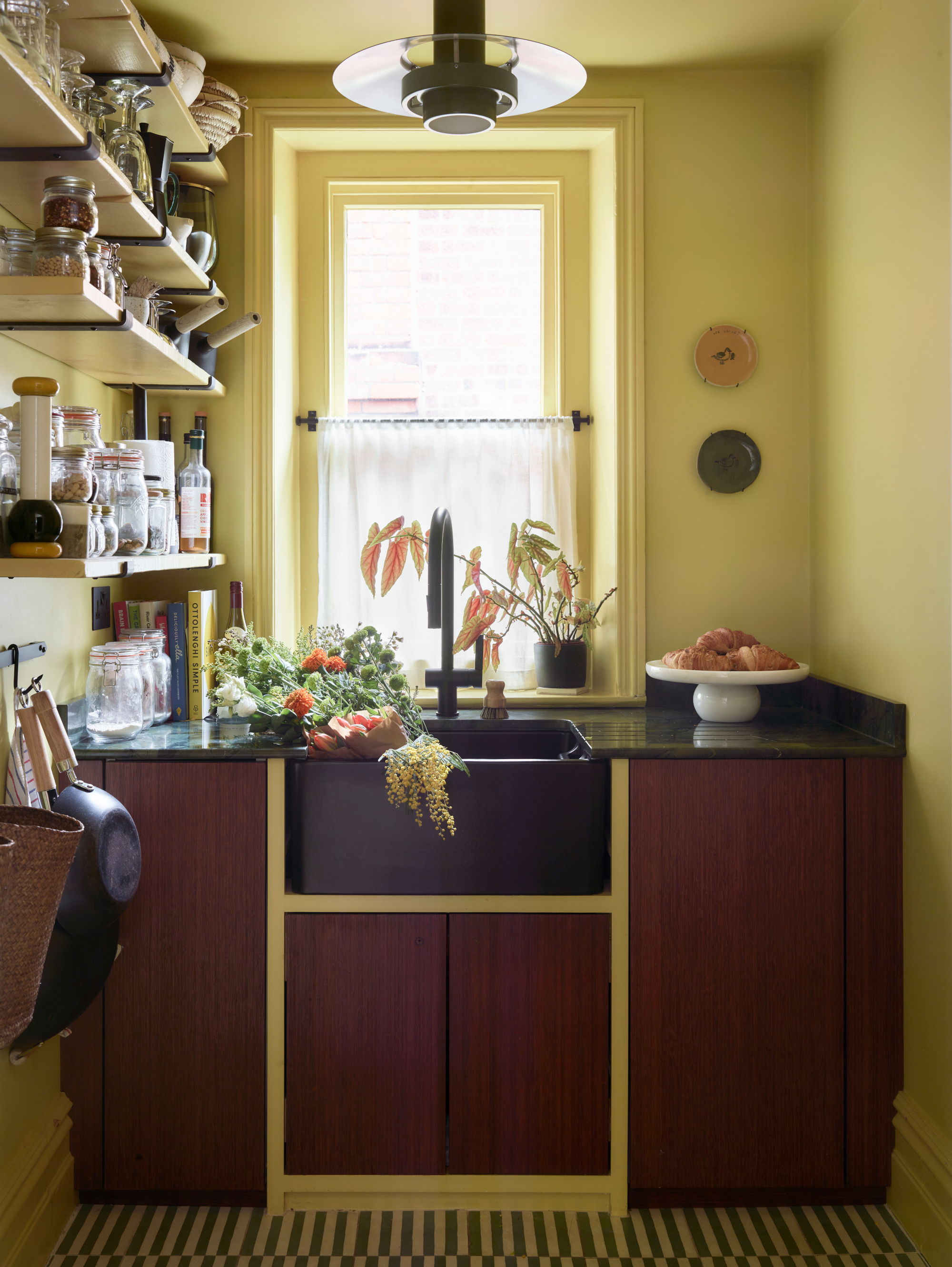Pantry room with dark red wood cabinets, black sink, shelves lined with food and black and white patterned floor tiles with yellow walls