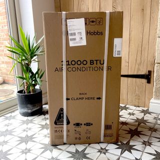 The Russell Hobbs RHPAC11001 Portable Air Conditioner in its brown packaging box on a tiled hallway floor